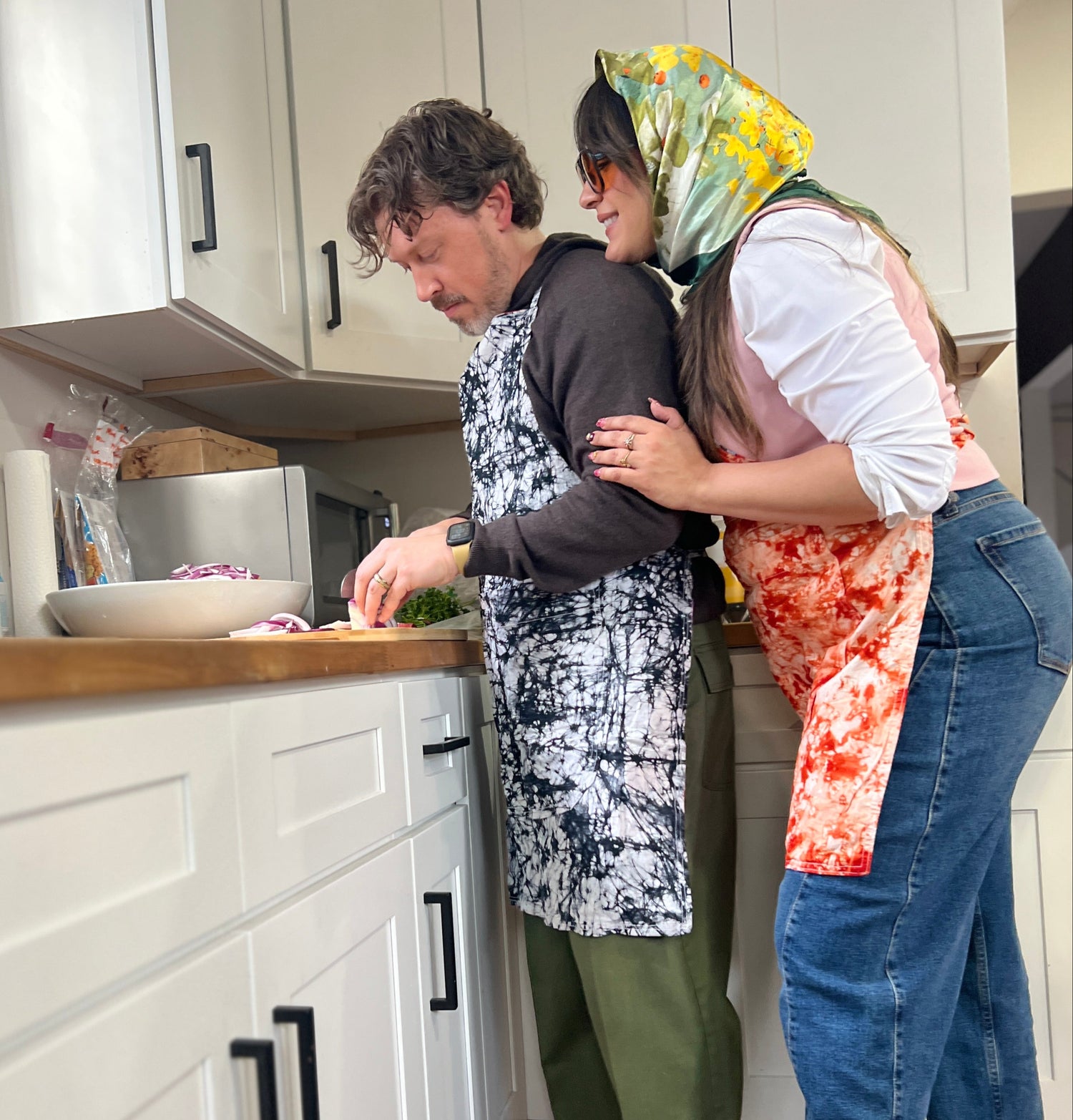 A couple in the kitchen wearing matching Echo aprons. The woman leans over the man’s shoulder as he preps a meal, sharing an intimate and joyful cooking moment.