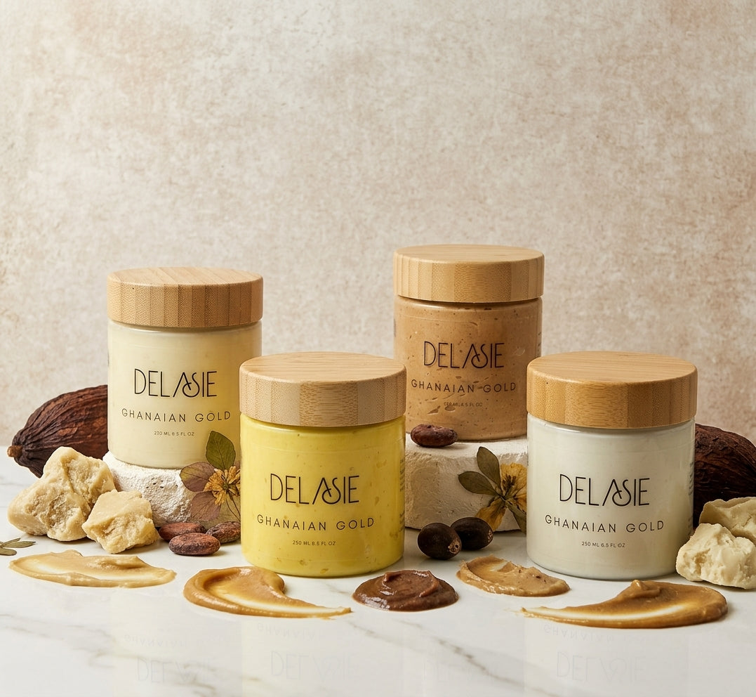 Four Delasie Ghanaian Gold cosmetic jars with bamboo lids arranged on a marble surface with cocoa beans, cocoa butter, and cream swirls. The jars contain cream-colored and yellow creams, set against a textured beige background.